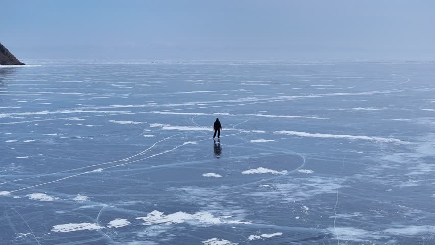 Cinematic tracking shot a lone ice skater on the vast, cracked crystalline surface of Lake Baikal under overcast skies.