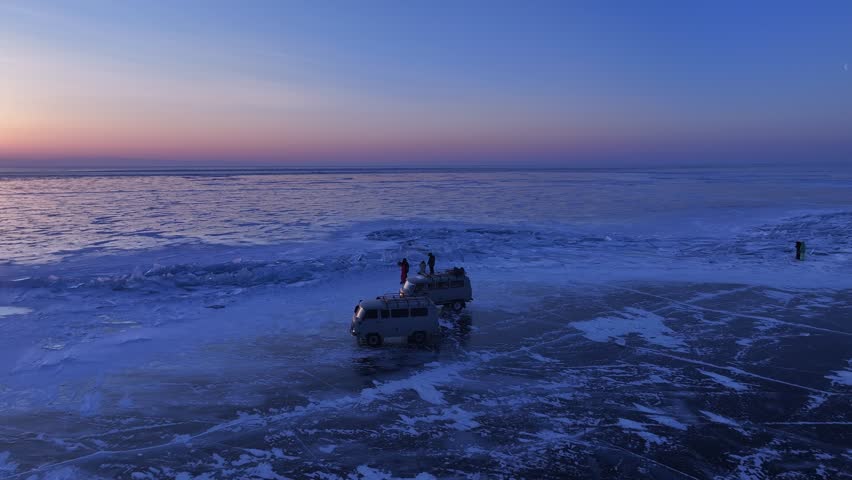 Serene orbit shot circling expedition vans and travelers on the crystalline frozen surface of Lake Baikal at twilight, 4K aerial.