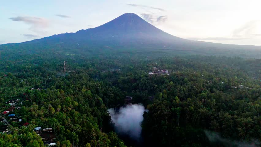 Cinematic 4K drone shot flying forward toward Tumpak Sewu waterfall and Mount Semeru volcano in soft morning light with mist rising from the lush green canyon in East Java Indonesia.