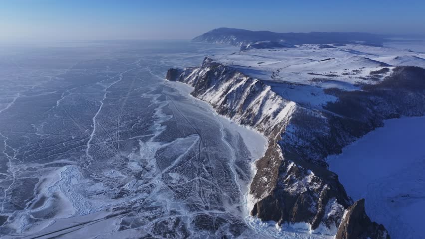 Majestic drone push in reveals the jagged snow-covered cliffs of Olkhon Island over the frozen, cracked surface of Lake Baikal at sunset.