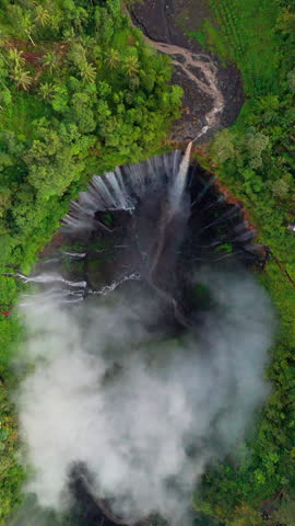 Cinematic 4K drone shot flying upward from a top down view over Tumpak Sewu waterfall surrounded by lush green rainforest and dramatic rising mist in East Java Indonesia.