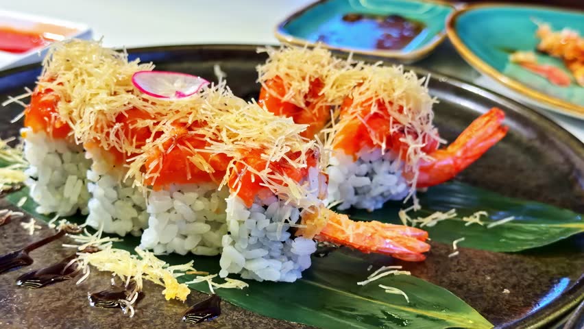 Close up footage of an ebi tempura sushi roll lifted with chopsticks at a restaurant table. Crispy shrimp, sushi rice, and sauce topping highlight fresh Japanese dining.