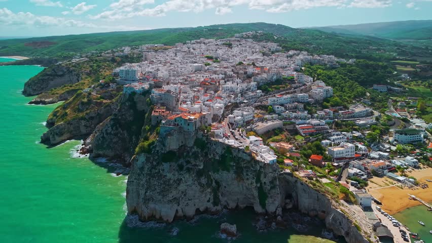 Aerial view of Peschici resort town on a sunny day in Puglia, Italy, popular Mediterranean destination with historic architecture, coastal cliffs, sandy beaches, and clear blue sea.