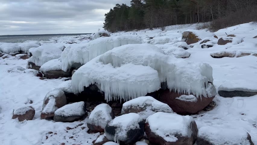 Slow panoramic shot of frozen rocky seashore with snow covered stones and icicles under dramatic cloudy winter sky, Baltic coast landscape.