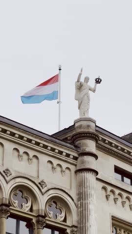 The national flag of Luxembourg waving next to a historic statue on the roof of a building