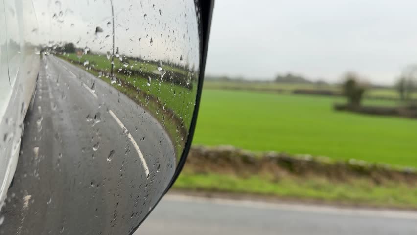 Reflection of cars driving on a wet rural road past a green field seen through a rain covered wing mirror