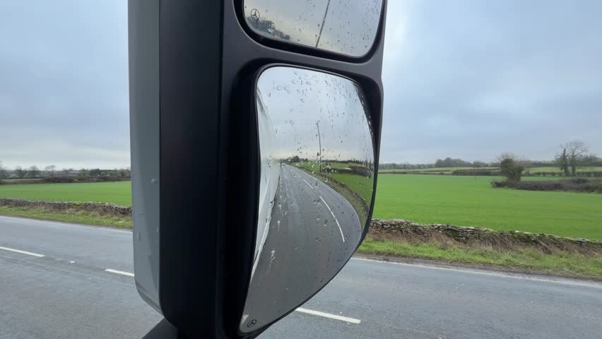 Reflection of cars driving on a wet rural road seen through a rain covered wing mirror past green fields