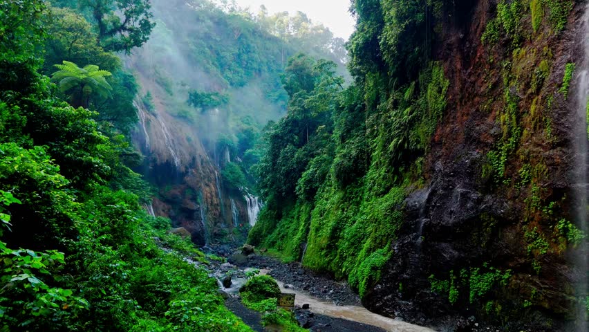 Drone flies through Tumpak Sewu canyon in Java, Indonesia, revealing lush green walls, mist, small waterfalls, winding riverbed, and serene tropical landscapes in 4K.