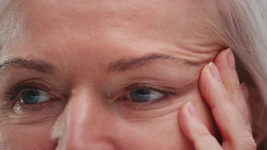 Close up: old woman eye. Middle aged female wrinkles near eyes. Aging, old skin, wrinkled face beauty portrait. Elderly lady touches skin on temples with fingers, facelift cosmetic cream. dermatology