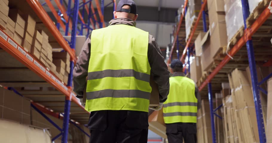 Warehouse workers carrying boxes between shelves. Men in vests move along racks, handling packages for delivery, shipping and inventory. Concept shows efficient warehouse order fulfillment.