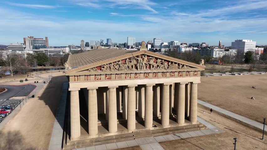 Rising aerial flyover of tNashville Parthenon in winter with downtown skyline in background under clear blue sky at Centennial Park, Tennessee. Tennis courts and calm pond in park. Sunny day. Rising.