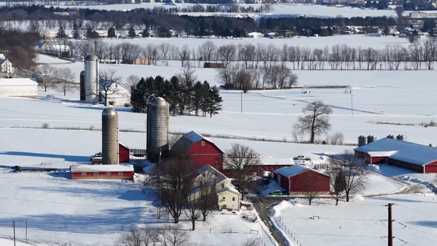 Aerial view of red barn and silos on snowy Pennsylvania farmland with farmhouse and rural fields during winter. Wide shot. Quiet and peaceful suburb district of town in United stages. Sunny day.