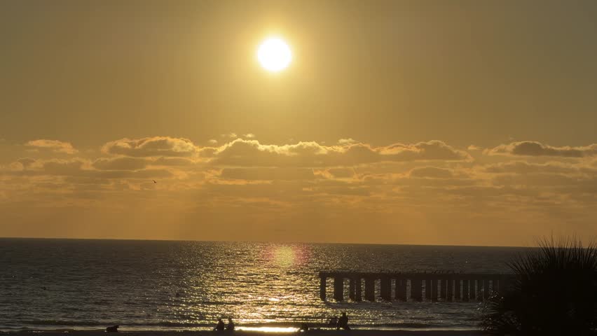 Golden sun illuminating the clear sky and ocean, reflecting on the water surface near a pier structure, creating a serene and warm atmosphere in South Florida