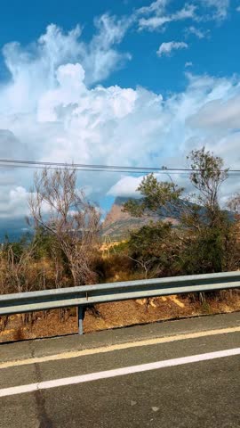 Roadside view of mountains under cloudy sky. Landscape shot from a road showing dry vegetation, trees, and a mountain peak partially covered by clouds.