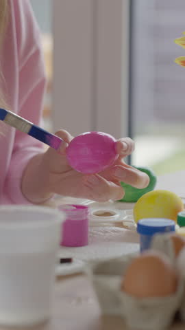 Woman painting Easter egg in pink color with brush at table with paints and other eggs. Concept of home leisure activity and preparation for spring holiday