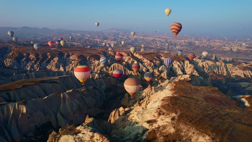 Hot air balloons floating and drifting across Cappadocia Turkey volcanic rock valleys at sunrise with aerial panoramic motion for travel and tourism promotion.