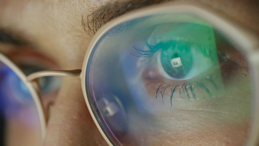 Extreme close up woman eye with glasses reflecting digital screen, browses internet, her pupils dilating, contracting, concentrates on online content. Woman eye with glasses reflecting computer screen
