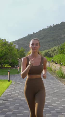 A fit woman in athletic wear jogs outdoors on a stone pathway surrounded by greenery and distant hills