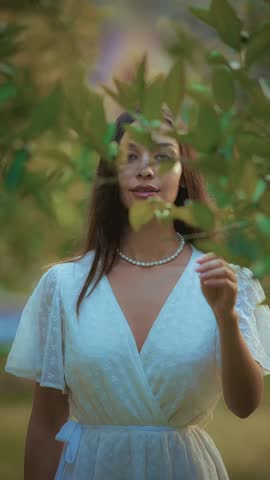 A beautiful young woman with long dark hair and blue eyes wears a delicate pearl necklace and a white textured dress while standing amidst vibrant green leaves in a natural setting