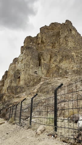 A shot of the historic Mount Uhud rocky cliffs and the sacred cave area in Medina. Features the steep desert terrain and protective fencing under an overcast sky at this significant Islamic heritage.
