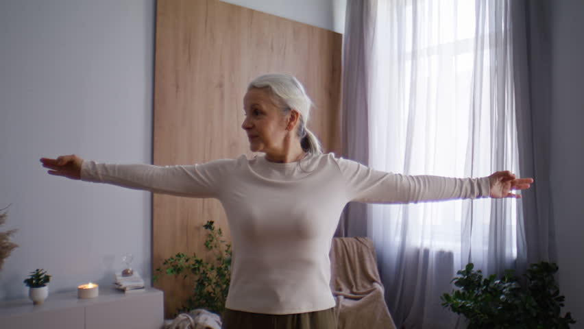 Elderly woman raises arms overhead practicing breathing exercise with gentle focus. Practitioner holds palms together inhaling deeply and exhaling slowly