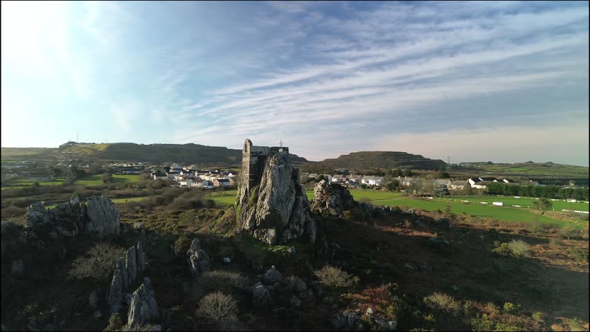 Smooth aerial drone footage orbiting around Roche Rock, a dramatic granite outcrop and historic chapel ruin located in Cornwall, United Kingdom. Filmed in ultra high definition 4K on a bright and clear winter morning, this cinematic shot showcases the rugged Cornish countryside, rolling hills, and distinctive rock formation under crisp natural light. Ideal for use in travel content, documentaries, historical features, nature films, and promotional visuals representing rural England and scenic landscapes.