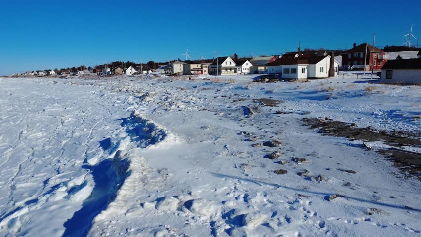 The winter shoreline of a small coastal village is covered in snow and large blocks of ice, under a brilliant blue sky. Baie-des-Sables, Gaspé Peninsula, Quebec, Canada.
