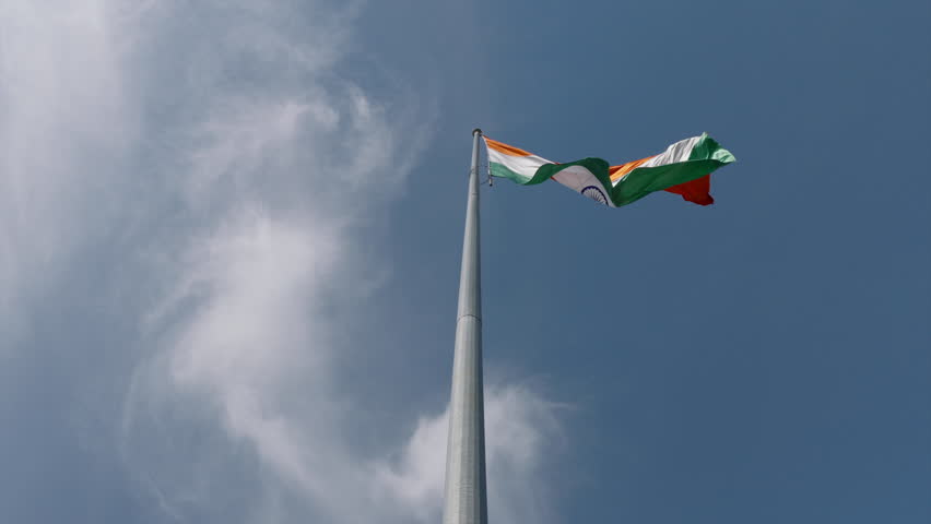 Low-angle slow motion shot of the Indian national flag waving against a blue sky with soft clouds, captured in 4K aerial footage.