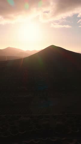 Aerial view shows La Geria planting pits, a lone white farmhouse, and a volcanic cone in Lanzarote as the camera moves downward and forward at golden hour.