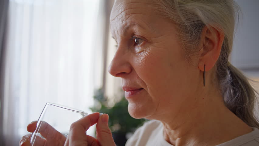 Senior woman takes painkiller with water sip in kitchen closeup. Elderly grey-haired lady uses medicine to reduce illness symptoms at home. Daily supplement