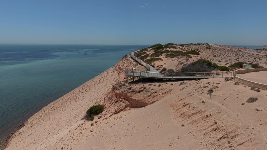 Aerial shot of a winding Eagle Bluff Lookout boardwalk to a sandy cliff lookout overlooking clear turquoise Waters of Henri Freycinet Harbour and an expansive arid coastline under a blue sky, Denham, Francois Peron NP, Shark Bay, Western Australia.