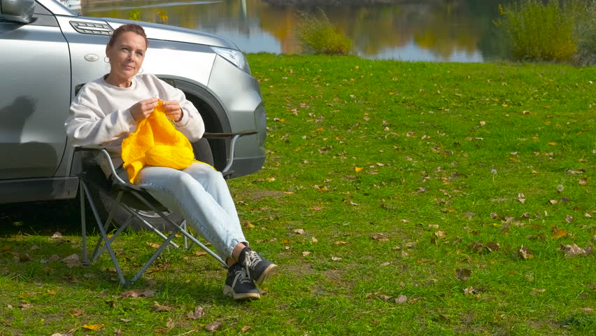 Relaxed woman knitting outdoors beside car. Woman sitting in a folding camping chair beside her car, peacefully knitting with bright yellow yarn while enjoying in nature by the lake during a road trip