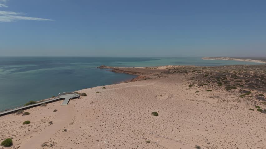 Aerial shot of a winding Eagle Bluff Lookout boardwalk to a sandy cliff lookout overlooking clear turquoise Waters of Henri Freycinet Harbour and an expansive arid coastline under a blue sky, Denham, Francois Peron NP, Shark Bay, Western Australia.
