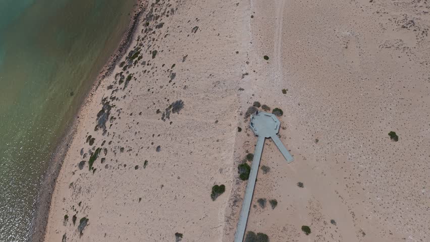 Aerial shot of a winding Eagle Bluff Lookout boardwalk to a sandy cliff lookout overlooking clear turquoise Waters of Henri Freycinet Harbour and an expansive arid coastline under a blue sky, Denham, Francois Peron NP, Shark Bay, Western Australia.