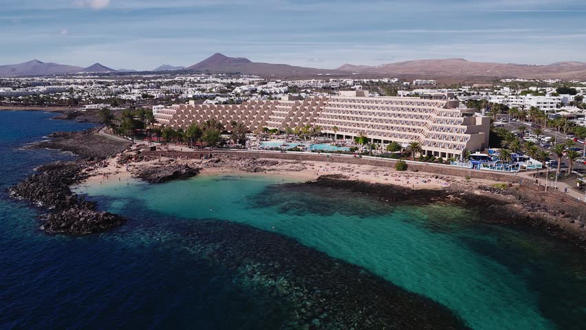 Aerial view shows a terraced hotel, pools, shallow lagoons, and sandy coves on Lanzarote as the camera glides, with beachgoers, lava rock, and brown volcanic hills.