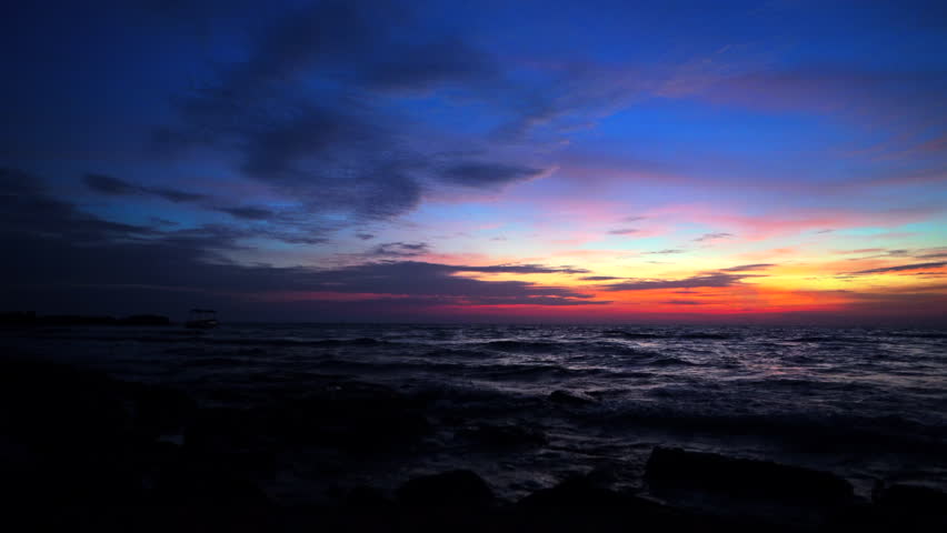 Sunset over ocean with boat in distance, rocks in foreground. Scene shows sunset over ocean with fishing boat visible in night. Rocks are scattered in foreground as waves crash gently. Sea landscape.