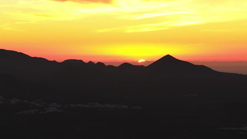 Elevated view shows the sun sinking behind a triangular cone and cratered peaks in Lanzarote, Canary Islands. Warm gradient backlights ridgelines and faint village buildings.