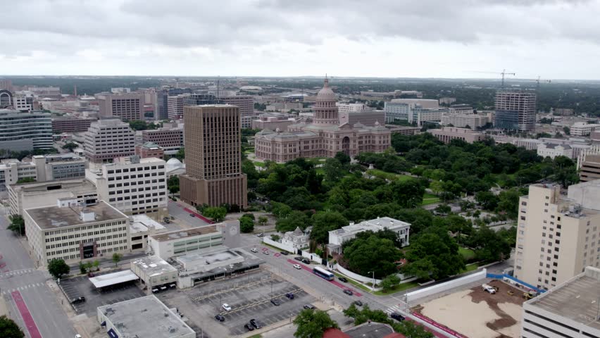 A sweeping aerial shot captures the iconic Texas State Capitol building nestled in lush green parkland, surrounded by the modern downtown Austin skyline. The sky is overcast, providing soft, diffused lighting over the cityscape, highlighting the contrast between historical architecture and contemporary high-rises.