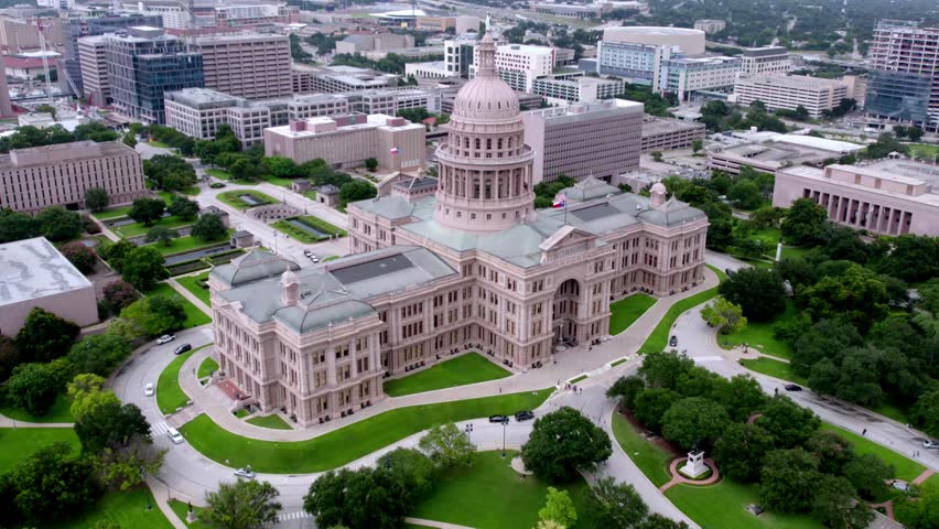An aerial, fly-through view showcasing the grand Texas State Capitol in Austin, Texas. The camera moves towards the dome, capturing the surrounding modern skyscrapers, manicured green lawns, pathways, and surrounding city streets under a daytime sky.