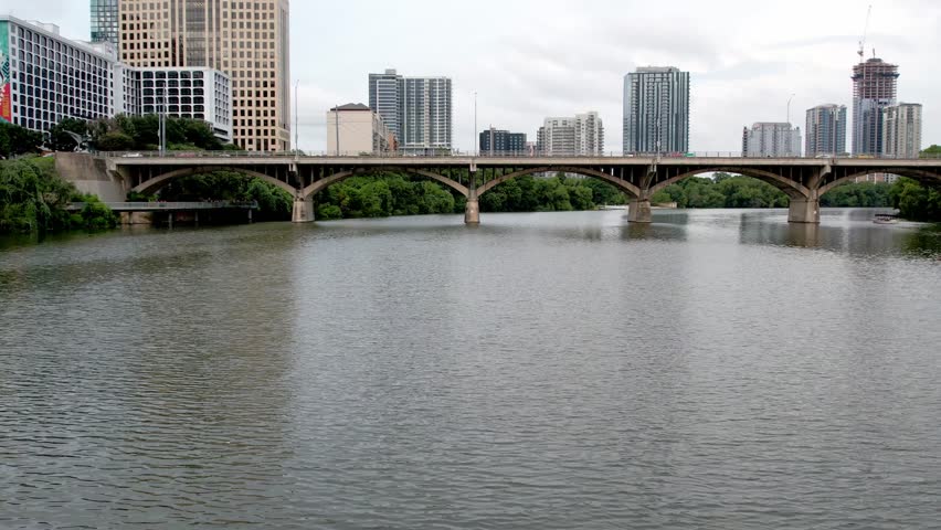 A sweeping, slightly elevated view of the Austin, Texas, skyline featuring modern high-rise buildings towering over lush green riverbanks. A prominent concrete arch bridge spans the wide, dark water of the Colorado River under an overcast, bright sky, suggesting a temperate urban landscape.