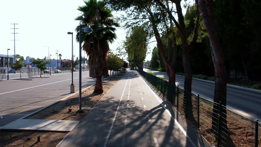 A forward-moving drone shot captures a dedicated bicycle path, marked with white lines, leading into the distance. Sunlight filters through tall trees creating strong shadows on the pavement. To the left is a parking area with sparse cars, and to the right, a metal fence separates the path from a multi-lane road with traffic.