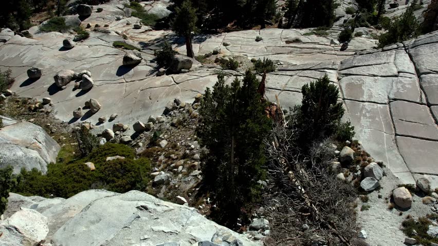 Aerial footage sweeping over a rugged, high-altitude landscape in Yosemite National Park. Massive, cracked granite domes and slabs dominate the foreground, interspersed with dark green pine forests and scattered boulders under a clear, bright blue sky. Distant mountains and iconic peaks frame the vast valley.