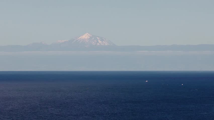 Aerial winter scene shows Gran Canaria cliffside harbor, white buildings, lighthouse, and boats leaving wakes, with Mount Teide rising above cloud on the horizon.
