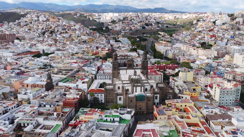 Aerial pan over Las Palmas rooftops and narrow streets centers on Catedral de Santa Ana, with Mount Teide on the horizon, soft daylight, cars and people move nearby.