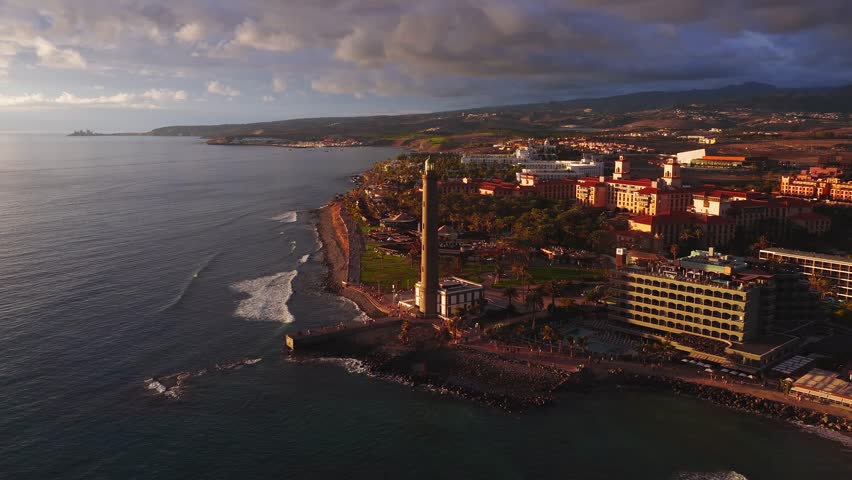 Aerial pan frames Faro de Maspalomas on a short pier in Gran Canaria, with palm promenade, hotels, rocky shore, people strolling, Atlantic waves, and distant mountains.
