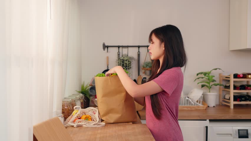 Asian young woman placing groceries on kitchen counter after shopping. Attractive beautiful girl preparing fresh fruits and vegetables, showing healthy lifestyle and home cooking routine in kitchen.