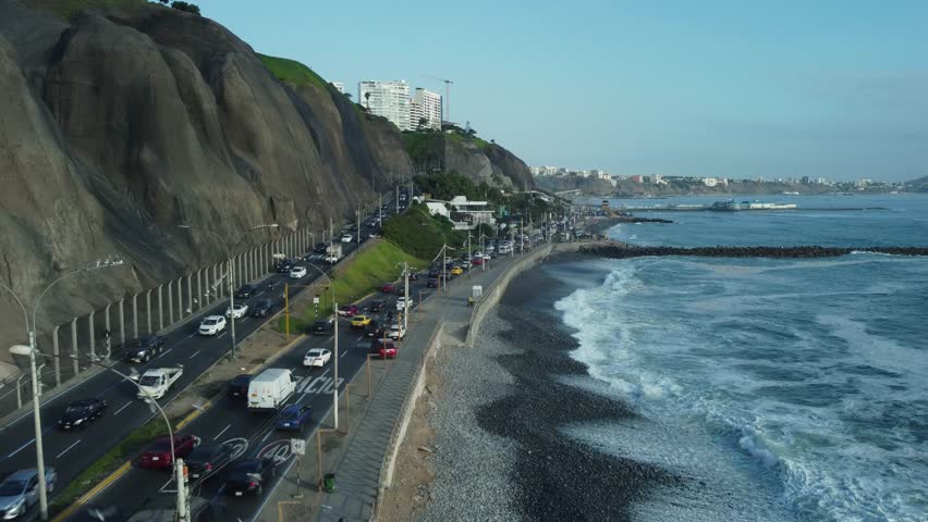 Aerial shot of a coastal highway named "Costa Verde". Drone flies forward as some birds fly around below and many cars drive on the road. Located in Miraflores district of Lima, Peru.