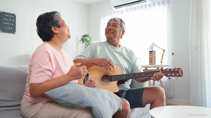 Asian elderly couple playing an acoustic guitar in living room at home. Loving senior older mature man and woman sitting on sofa, enjoying retirement life together share joyful bonding moment in house