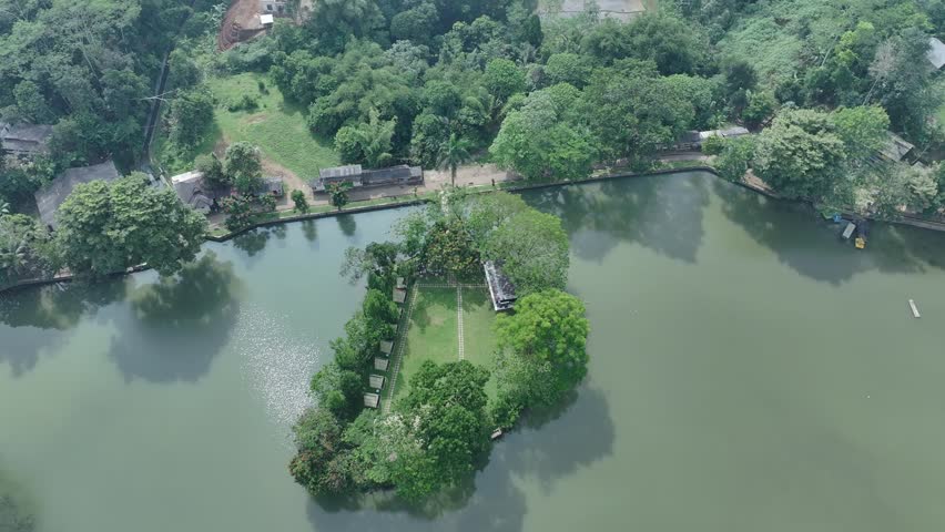 An aerial view of a small tree-covered island in the middle of a calm lake, surrounded by lush green landscape and rural settlements. The scenic natural environment features clear water, dense vegetation, and bright daylight conditions.