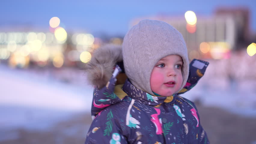 A warmly bundled child stands outdoors, winter lights glowing softly in the blurred background
